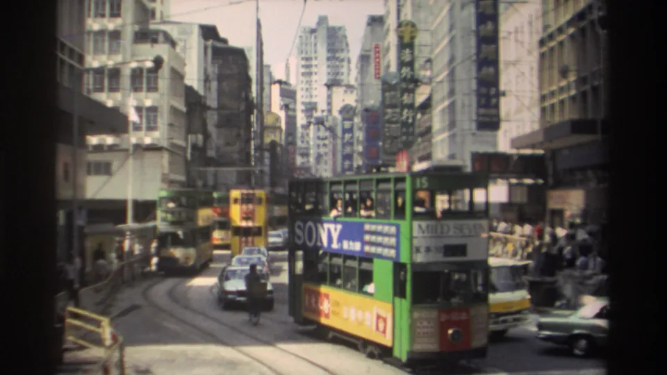 1982 Hong Kong traffic scene with a green tram turning along tracks, cars close by and pedestrians along the sidewalk.