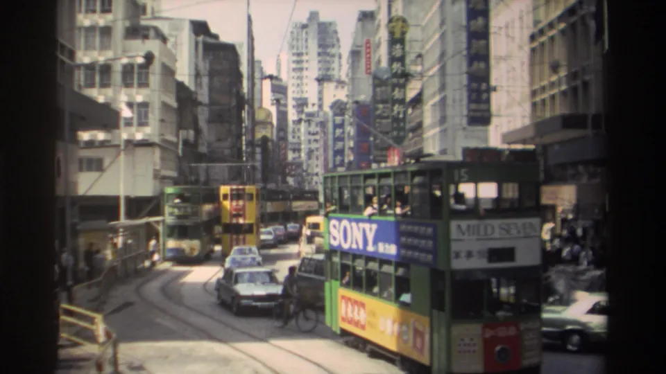 Green double-decker streetcar in 1982 Hong Kong passing cars, cyclists or pedestrians, shop signs and dense buildings.