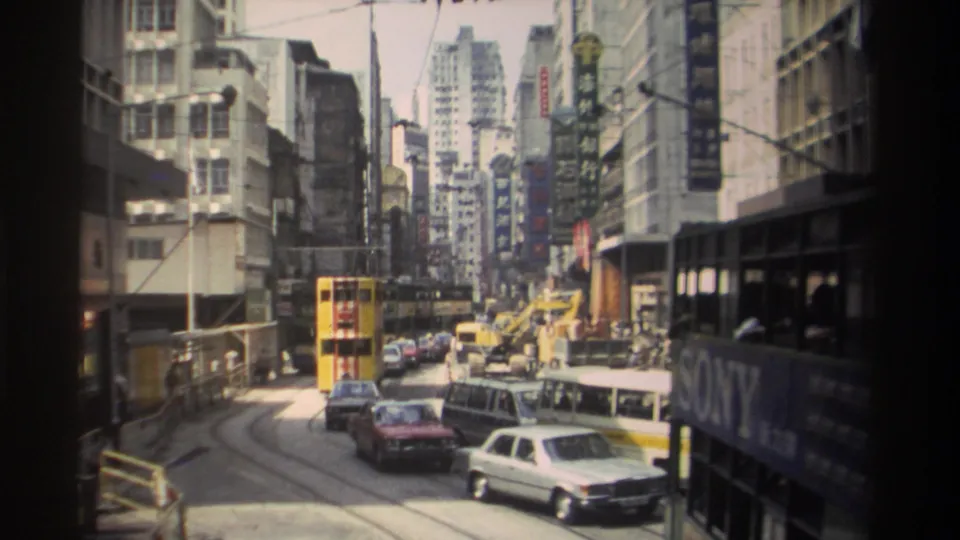 1982 Hong Kong street footage with a yellow tram, red car, white vehicle, construction vehicle, tram tracks, and dense building signage.