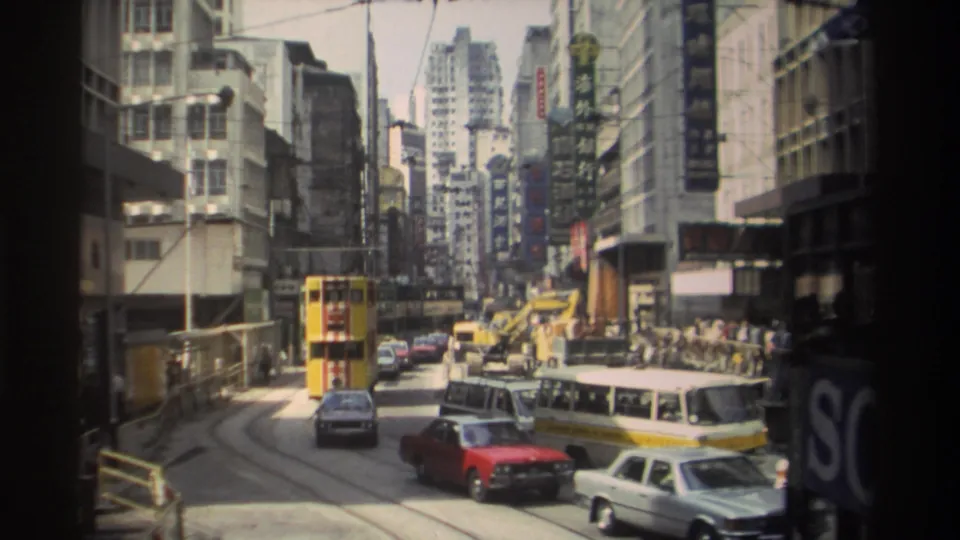 1982 Hong Kong street view with trams, classic cars, a white van, construction machinery, pedestrians, and Chinese signage along the buildings.
