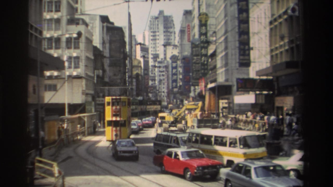 1982 Hong Kong street view with a yellow double-decker bus, red and silver cars, tram tracks, tall buildings, and sidewalk pedestrians.