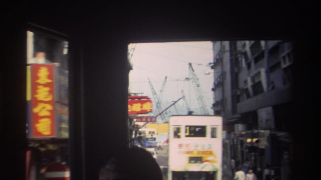 Hong Kong street traffic seen through a dark framed foreground, with a bus, Chinese signage, pedestrians, and construction cranes beyond.
