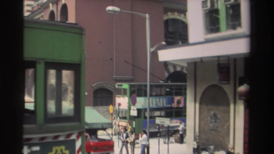 1982 Hong Kong street corner with a green tram or tramcar, red car, pedestrians, shopfronts, and dense mid-rise facades.