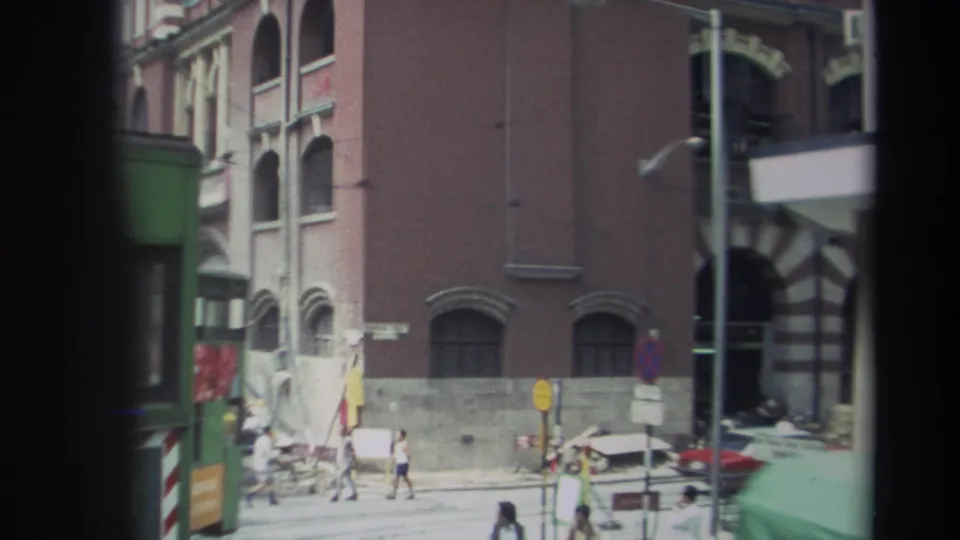 1982 Hong Kong street corner seen from a moving vehicle, with pedestrians, traffic signs, brick walls, and a green vehicle edge.