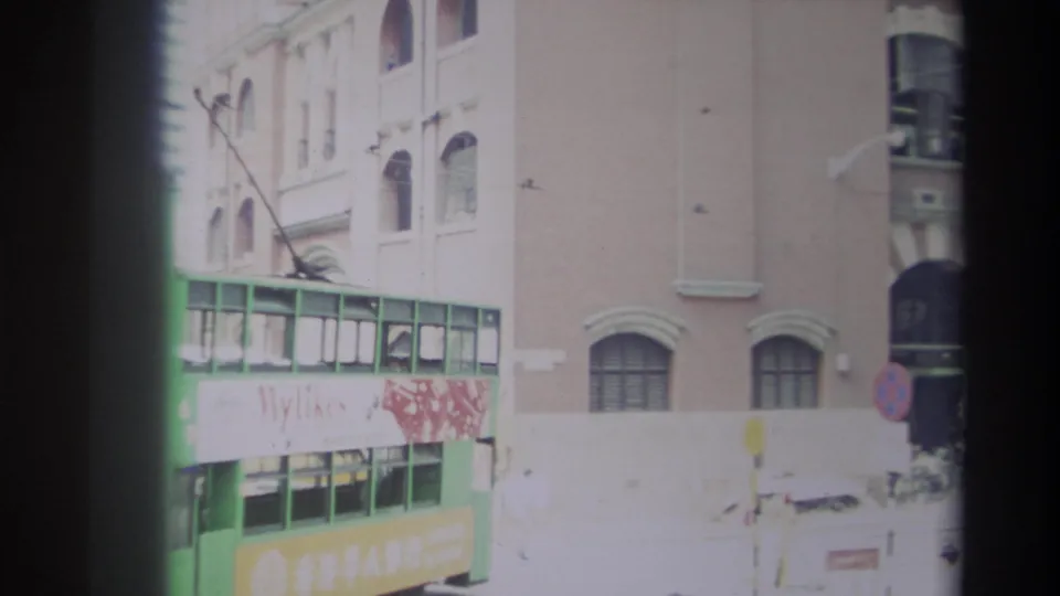 Hong Kong 1982 street view with a green trolley or tram-like vehicle, faded signage, brick facade, and passing traffic.