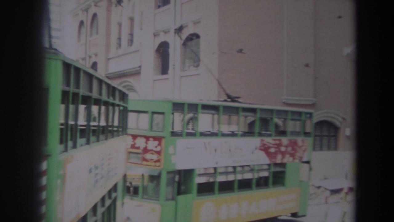 Green public transit vehicle in 1982 Hong Kong turns through a narrow street with signage, wires and masonry facades visible.