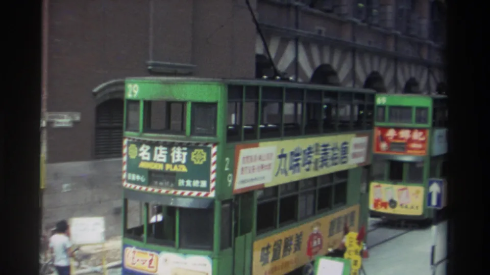 Elevated 1982 Hong Kong view of two green double-decker trams, route signage, advertising panels, and pedestrians near the curb.