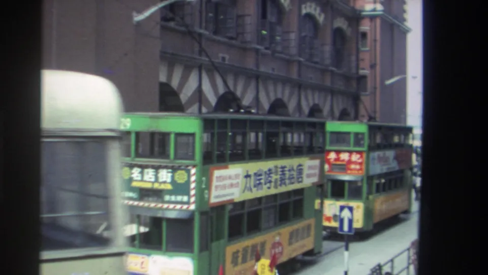 A white bus partially blocks the foreground as two green double-decker trams continue along a Hong Kong street in 1982.
