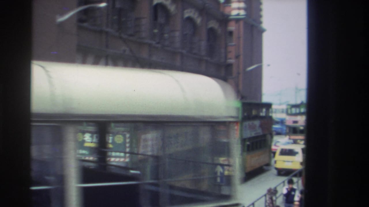 1982 Hong Kong street view with a green double-decker bus moving past a covered structure and older brick facades.