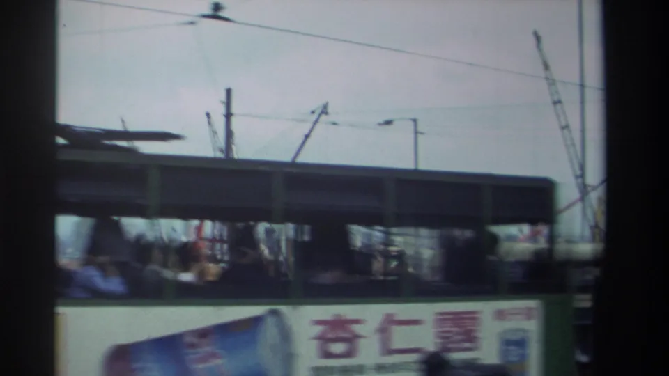 A double-decker bus in 1982 Hong Kong moves past the camera with passengers, Chinese signage, wires, and cranes behind it.