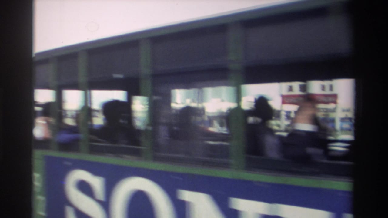 Side of a double-decker bus with large lettering and passengers visible through windows.