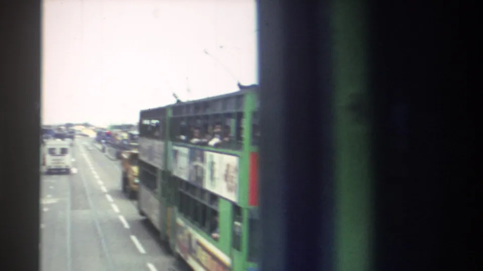 Moving 8mm view along a Hong Kong street, partly blocked by a vehicle-window edge, with bus traffic and road markings below.