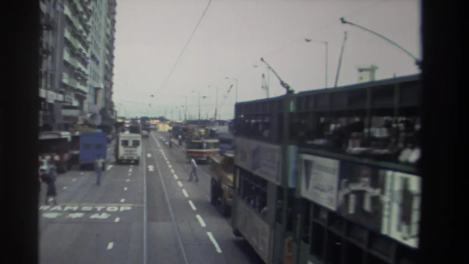 Wider 1982 Hong Kong traffic view with double-decker buses, cars, road markings, a crossing pedestrian, and tall buildings.