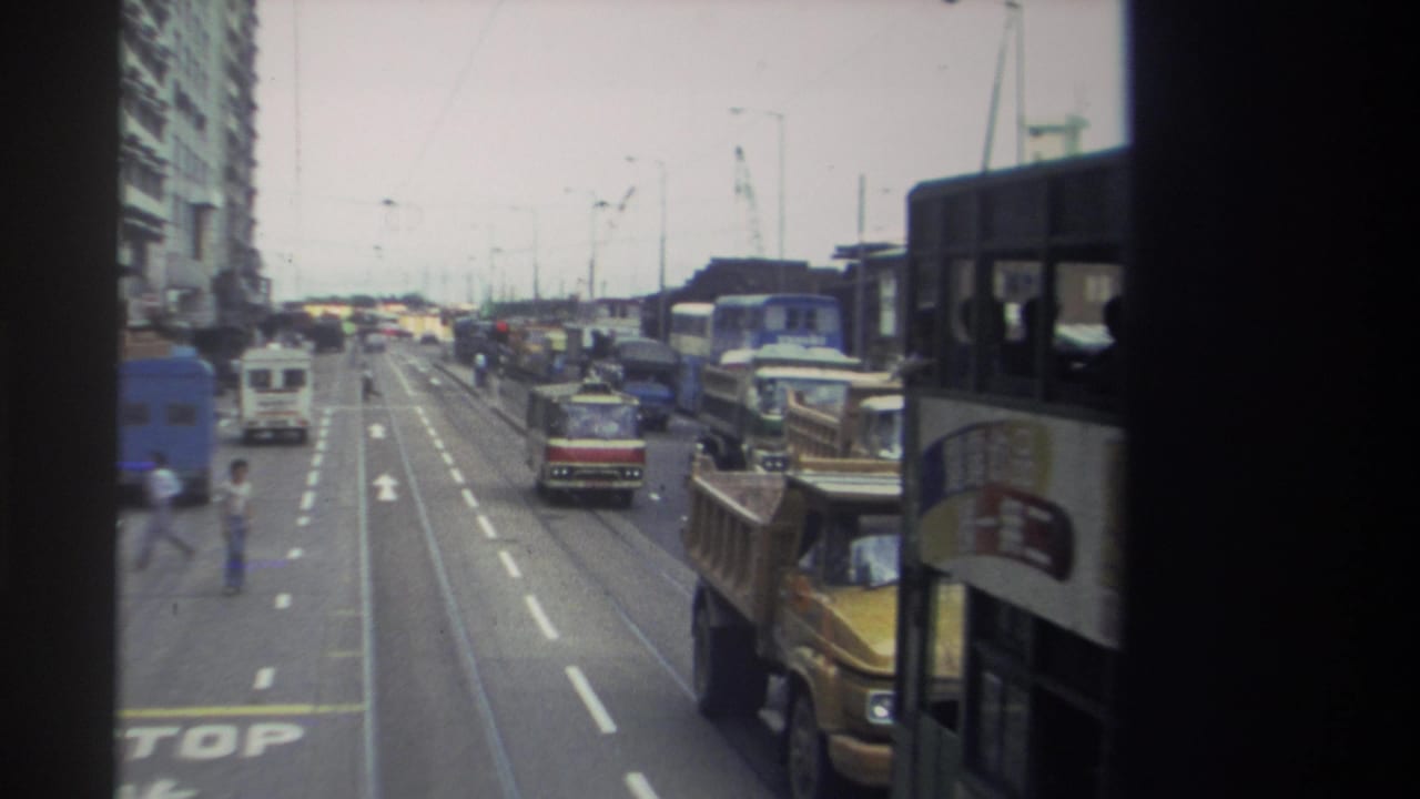 Hong Kong street traffic in 1982 with double-decker buses, a truck, vans, tram tracks, overhead wires, and tall roadside buildings.