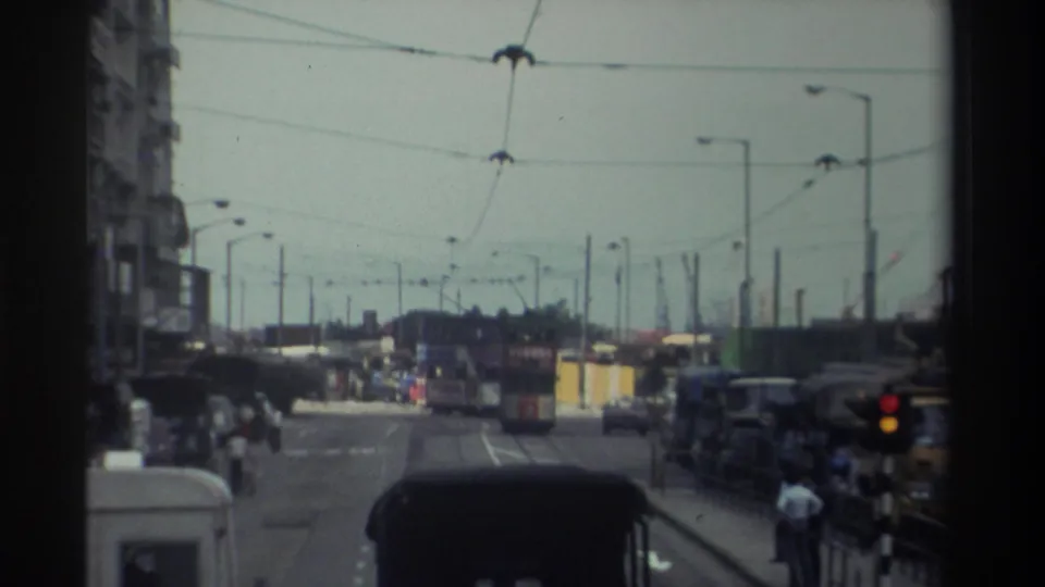 Moving-vehicle view of a Hong Kong roadway in 1982 with overhead tram wires, traffic lights, buses, cars, pedestrians, and roadside structures.