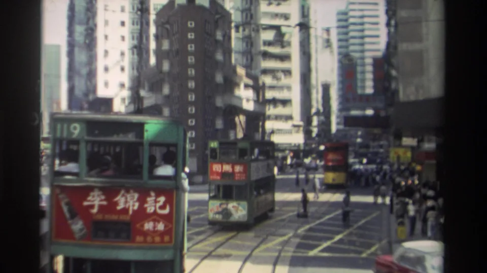1982 Hong Kong traffic scene with a route 119 tram in front, another tram ahead, pedestrians crossing, and high-rise buildings around the tracks.