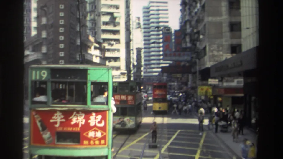1982 Hong Kong intersection view with a green double-decker tram, pedestrians crossing the tracks, a yellow tram ahead, and dense buildings lining the street.