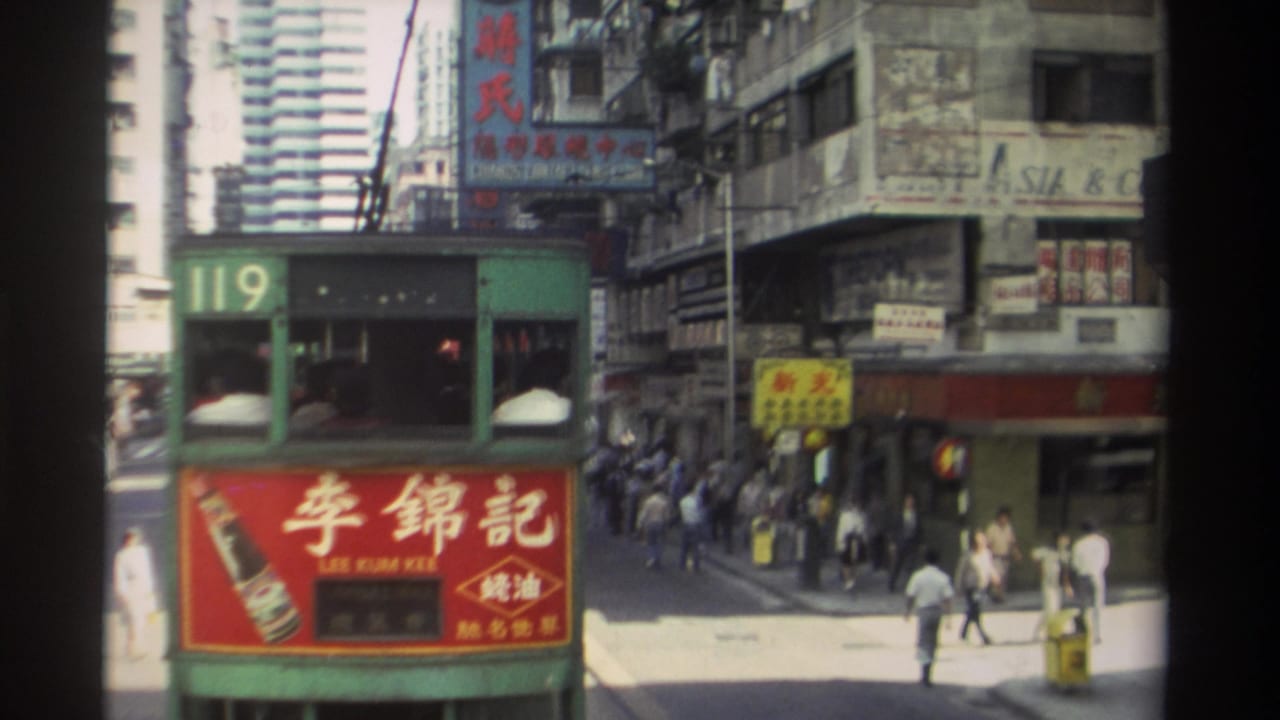Green Hong Kong tram 119 moves through a sunny street scene with Chinese signage, pedestrians, shopfronts, and tall buildings behind it.
