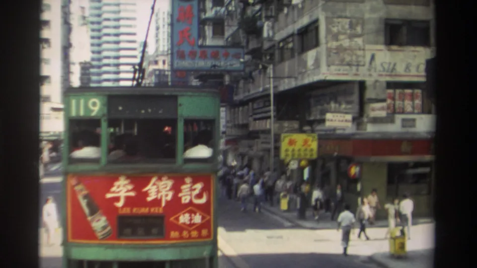 Green Hong Kong tram 119 moves through a sunny street scene with Chinese signage, pedestrians, shopfronts, and tall buildings behind it.
