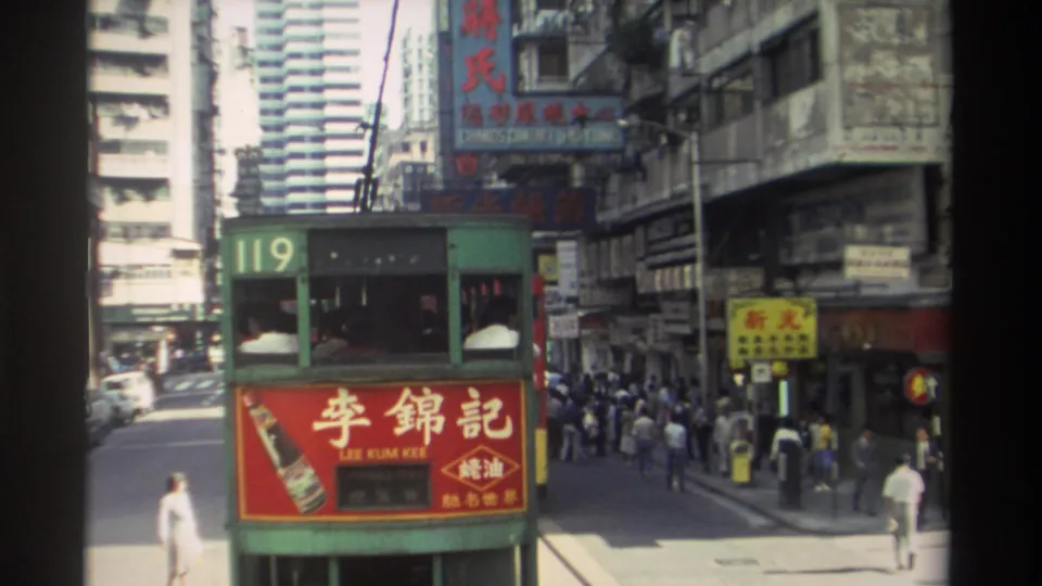 1982 Hong Kong street frame showing tram 119, a red Lee Kum Kee panel, pedestrians, road markings, and layered Chinese commercial signs.