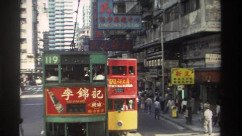 1982 Hong Kong traffic scene with green tram 119, a red double-decker bus, pedestrians, Chinese signage, and commercial storefronts.
