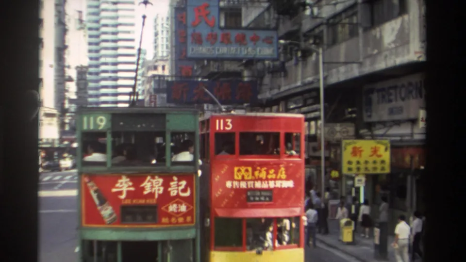 1982 Hong Kong street view with green and red double-decker trams, shop signs, pedestrians, and tall buildings in the background.