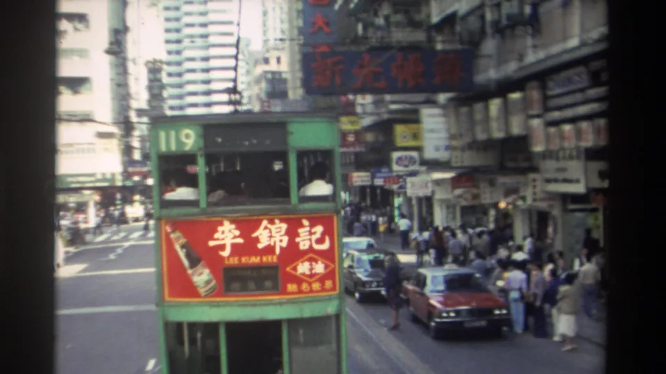 1982 Hong Kong street scene with a green tram, pedestrians waiting by storefronts, cars at the curb, and layered commercial signs.