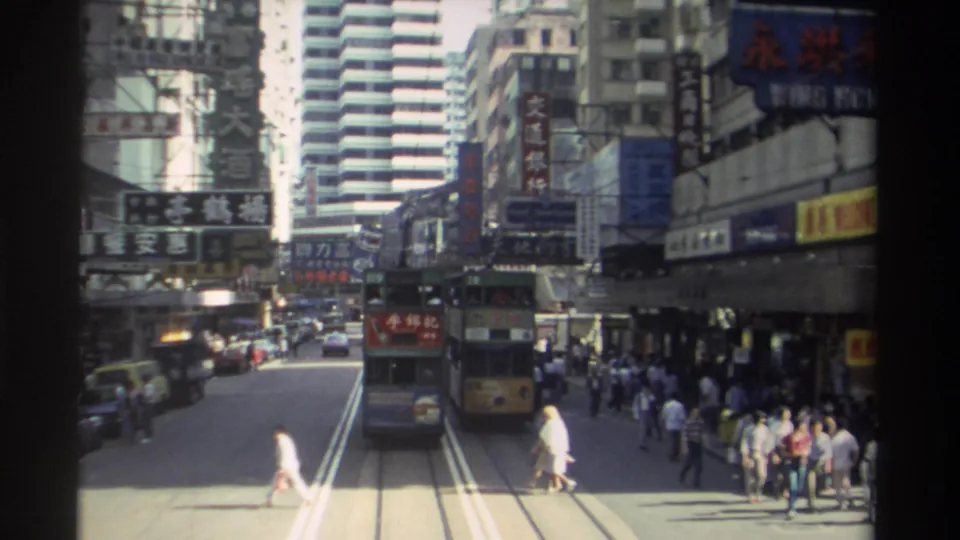 Wide 1982 Hong Kong street view showing tram tracks, double-decker trams, pedestrians crossing, storefront signage, and high-rise buildings.