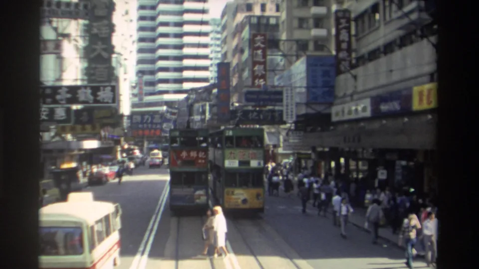 1982 Hong Kong traffic scene showing a tram moving down the tracks, a cream minibus in the foreground, pedestrians along the sidewalk, and layered Chinese commercial signs.