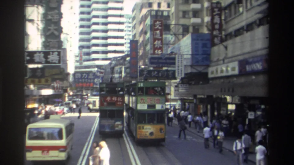 1982 Hong Kong street scene with a double-decker tram approaching beside a cream minibus, pedestrians crossing and walking near storefronts, and Chinese signs over the roadway.