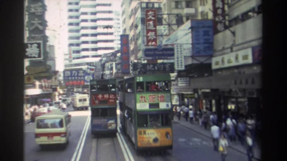 1982 Hong Kong urban traffic view with a green and yellow double-decker tram, a cream minibus, pedestrians under shaded storefronts, tall buildings, and prominent Chinese signage.