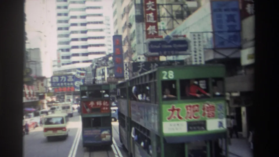 Close 1982 Hong Kong street view of tram number 28 beside cars, pedestrians, shop signs, and tall commercial buildings.