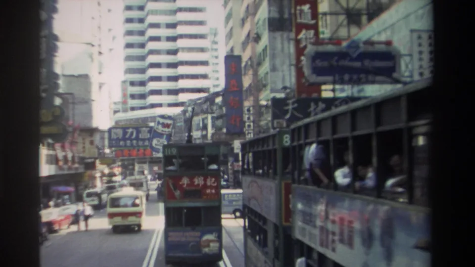 1982 Hong Kong traffic scene with double-decker trams, a cream car, pedestrians, vertical signs, and dense commercial street frontage.