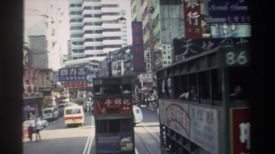 1982 Hong Kong street footage showing a close passing tram, another tram ahead, cars, pedestrians, and layered Chinese signage.