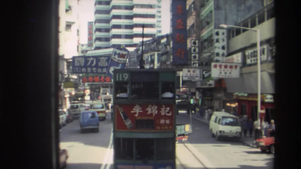 1982 Hong Kong tram street scene with the route 119 tram centered, compact vehicles nearby, pedestrians at the curb, and dense commercial signage.