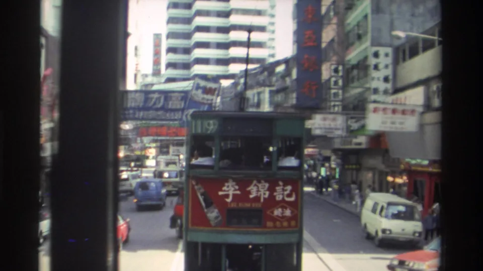 A 1982 Hong Kong double-decker tram with a Lee Kum Kee advertisement moves through traffic beneath Chinese-character shop signs.