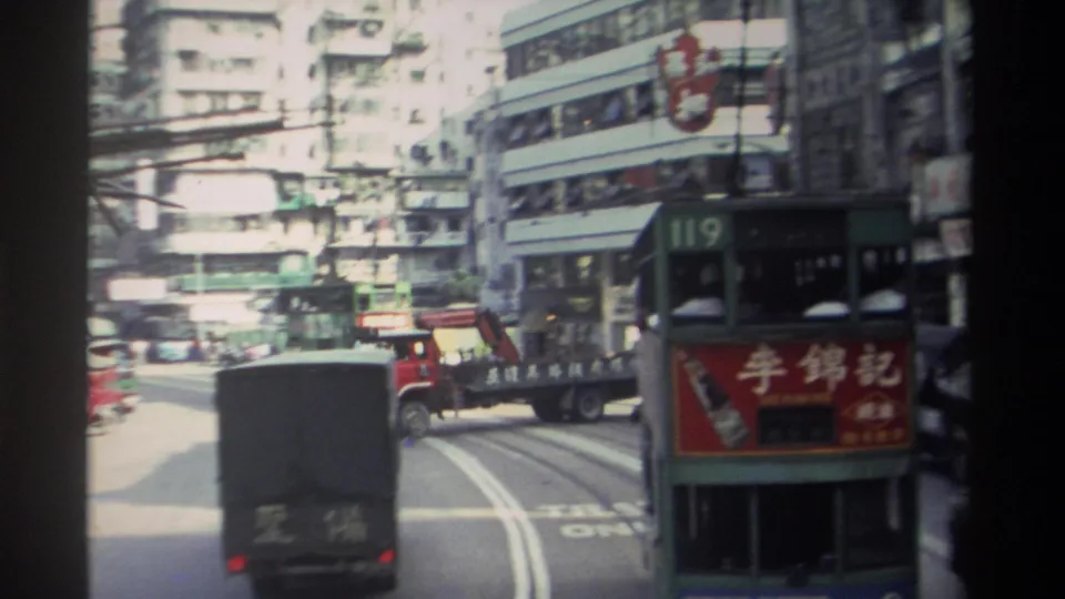 1982 Hong Kong traffic view with a box truck, a green route 119 double-decker tram, tram tracks, and shopfront signs.