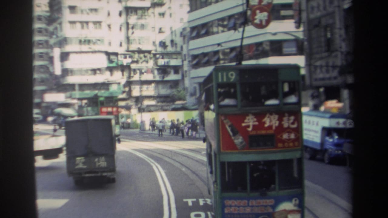 1982 Hong Kong street view from a moving vehicle with a green double-decker tram, delivery truck, pedestrians, and dense buildings.