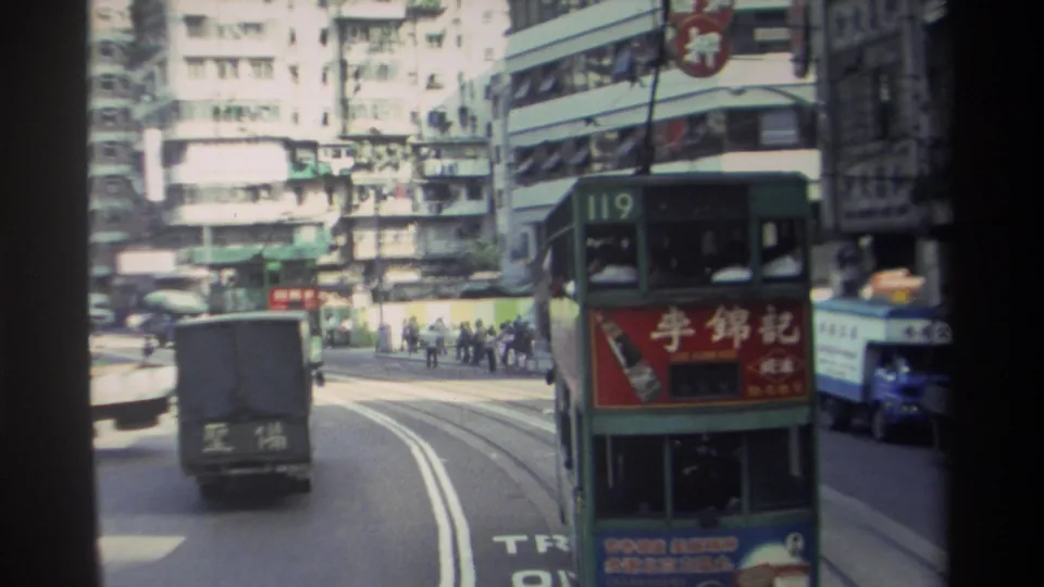 1982 Hong Kong street view from a moving vehicle with a green double-decker tram, delivery truck, pedestrians, and dense buildings.