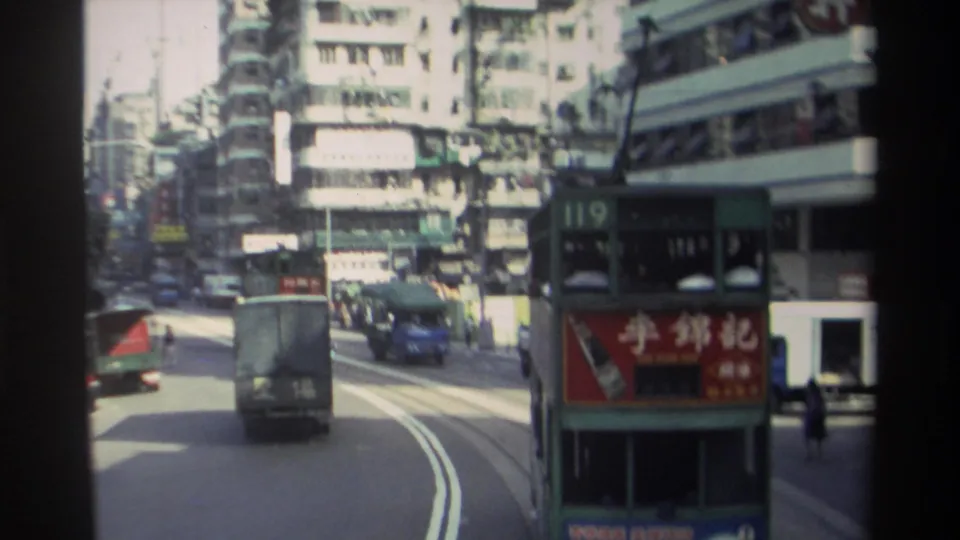 Moving 8mm view of a Hong Kong street in 1982, showing a tram, delivery truck, blue vehicle, sidewalks, and tall buildings.
