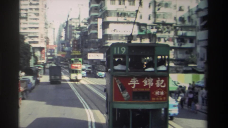 Closer view of route-119 Hong Kong tram with red brand advertisement, tram tracks visible in the road surface, dense urban architecture and Chinese signage surrounding, 1982 8mm film.