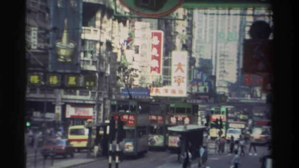Wide view of a dense Hong Kong commercial street circa 1982, with Chinese and bilingual storefront signs, a tram, pedestrians, overhead wires, and packed mid-rise buildings.