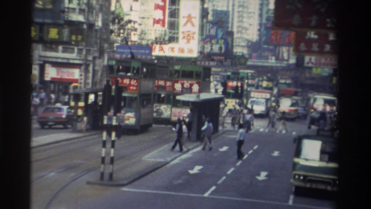 Pedestrians cross a busy Hong Kong street with a double-decker tram and Chinese commercial signage on high-rise buildings behind.