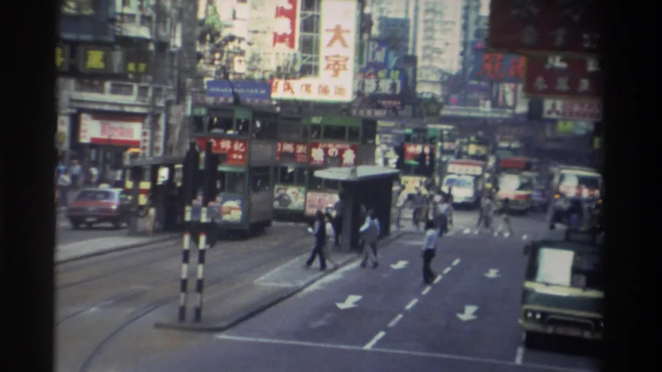 8mm footage, 1982 Hong Kong — pedestrians at a marked crosswalk, green double-decker tram in mid-ground, buildings plastered with Chinese-character signs in red and yellow.