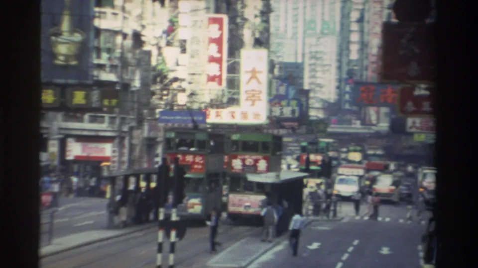 1982 Hong Kong, 8mm — tram and vehicles on a multi-lane street; buildings behind are densely covered with Chinese-character commercial signs, one large white sign prominent in frame.