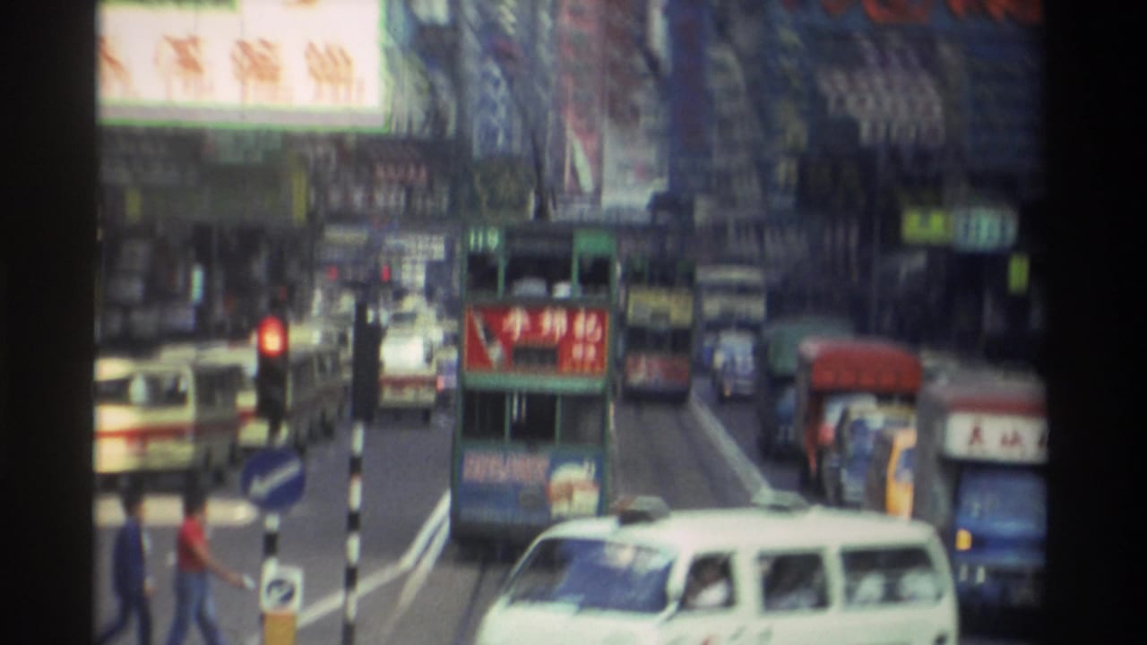 Overhead view of a busy Hong Kong commercial street circa 1982, showing a green double-decker tram, white minibus, red traffic light, and buildings covered in Chinese billboard signs.