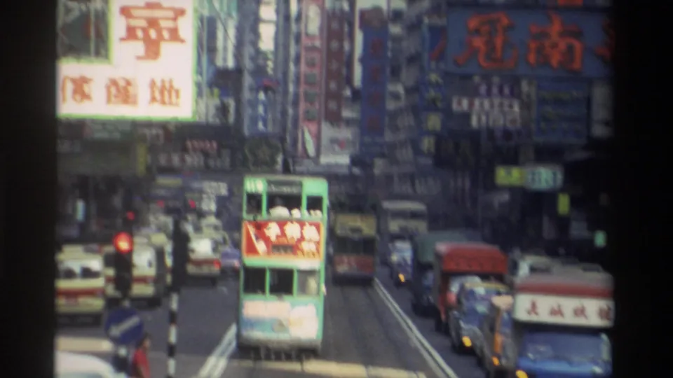 1982 Hong Kong busy street from elevated vantage, with green double-decker tram, white vehicles, red traffic signal, and large Chinese-character billboard signs on surrounding buildings.