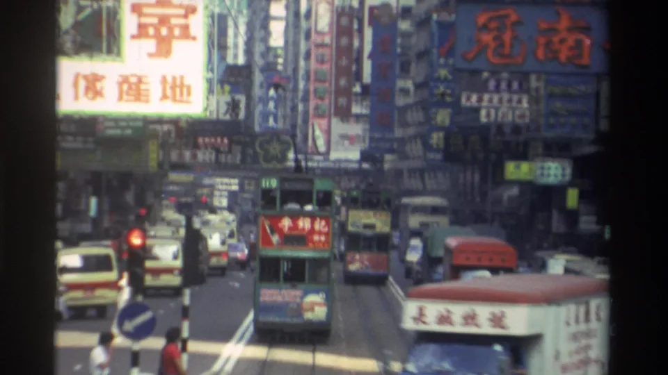 1982 Hong Kong commercial street from above, with prominent Chinese-character billboards, green double-decker tram, white minibus, red traffic light, and pedestrians in the foreground.