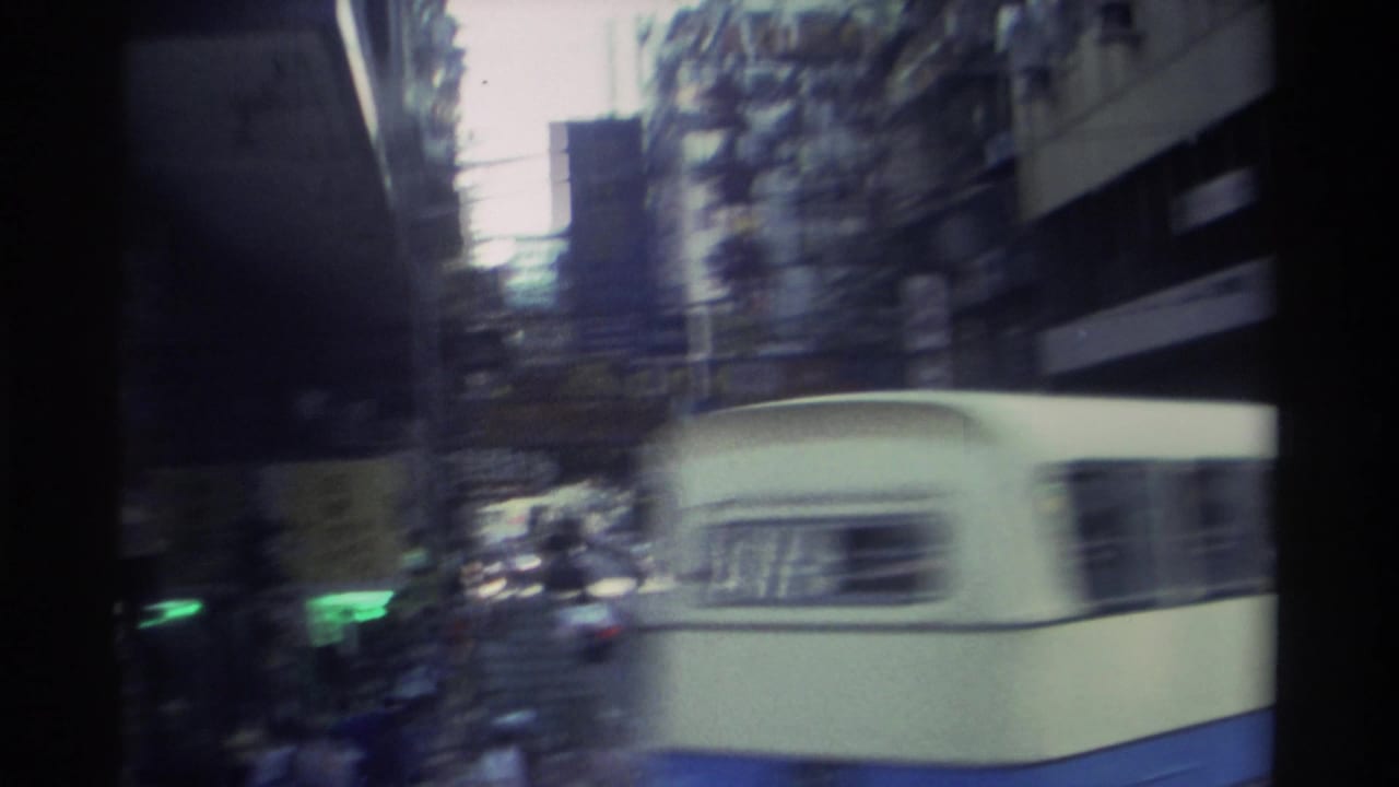 White double-decker bus blurs past the 8mm camera in 1982 Hong Kong; tall buildings and street infrastructure are visible through the motion.