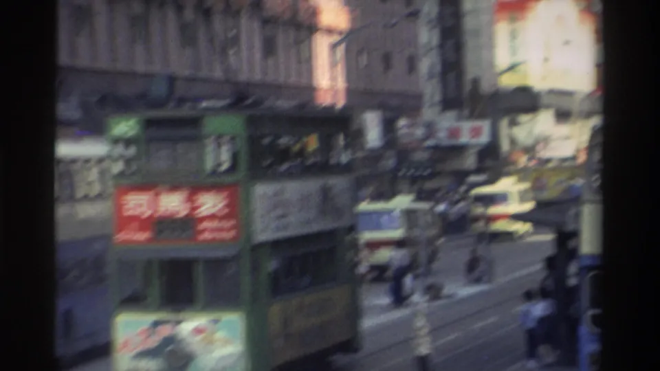 Hong Kong street corridor in 1982 as seen from a moving vehicle: green trams, dense Chinese shopfront signage, and a Coca-Cola advertisement on a building facade.